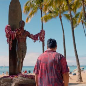 man in red short sleeve button down stands underneath a statue on the beach with palm trees