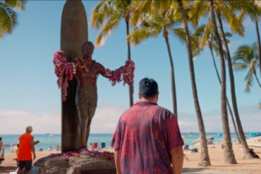 man in red short sleeve button down stands underneath a statue on the beach with palm trees