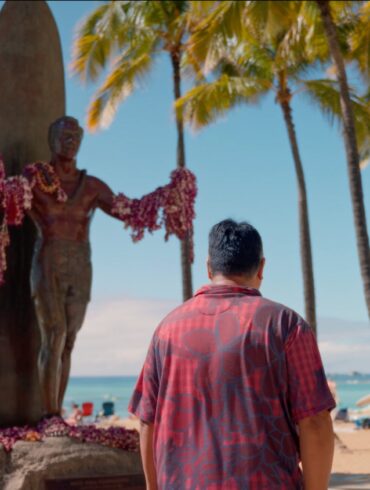 man in red short sleeve button down stands underneath a statue on the beach with palm trees